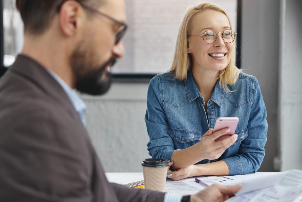 Two intelligent female and male office managers meet together for doing common task, surrounded with many documents and takeaway coffee, use modern electronic device for work and communication
